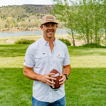 Man standing outdoors holding a football wearing a white shirt called The Stay Late and a tan cap