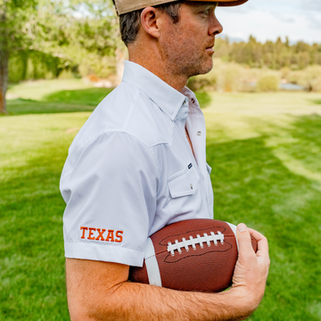Man wearing "The Stay Late" white shirt holding a football on a grassy field with trees in the background.