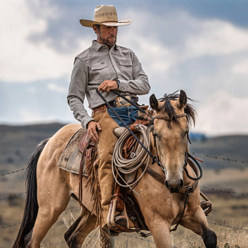 Man wearing The Stockton shirt and cowboy hat riding a horse in an open landscape with cloudy sky