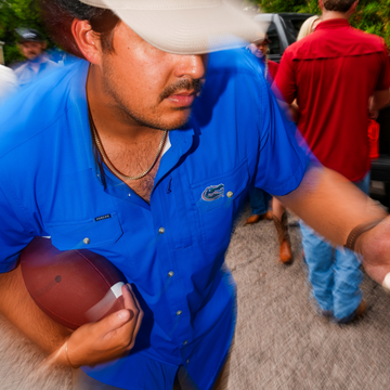 Man wearing bright blue shirt holding football outdoors with group in background, featuring The Swamp Chomp design