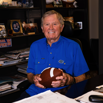 Older man in a blue button-down shirt holding a football at a desk in a sports memorabilia office