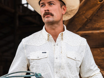Man wearing a white patterned shirt called The Vega and a cowboy hat, holding a rope outdoors