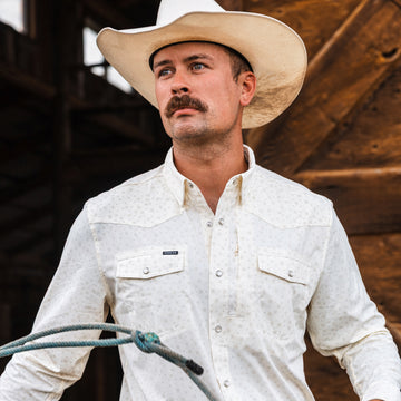 Man wearing a white patterned shirt called The Vega and a cowboy hat, holding a rope outdoors