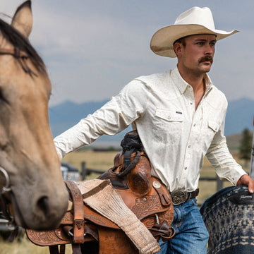 Man wearing The Vega shirt and a white cowboy hat holding a saddle near a horse in an outdoor setting