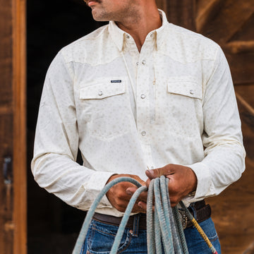 Man wearing The Vega long-sleeve shirt holding coiled rope in a rustic wooden setting outdoors