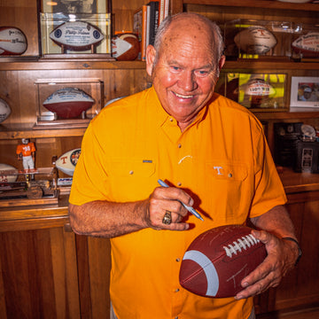 Smiling man in an orange shirt holding a football and pen in front of sports memorabilia shelves