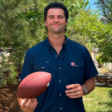 Man smiling outdoors while holding a football and wearing a navy button-up shirt with chest pockets