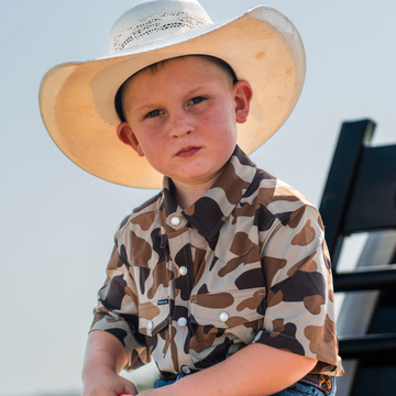 Young boy wearing a wide-brimmed hat and camo shirt sitting outdoors, showcasing The Wingman shirt.