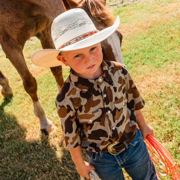 Young boy wearing The Wingman shirt with cowboy hat standing next to a horse holding a rope outdoors