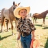 Young boy wearing The Wingman shirt and cowboy hat standing outdoors near horses holding a rope rope