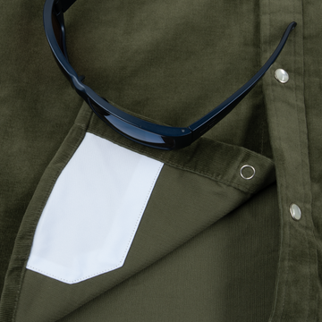 Close-up of olive green fabric with white inner pocket detail and black sunglasses resting on The Zilker shirt.