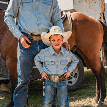 Young cowboy standing next to his dad in a light denim shirt