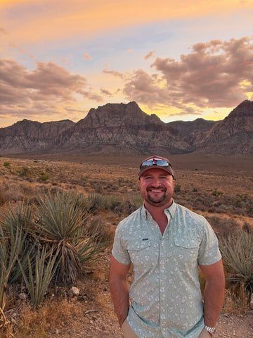 Man wearing a light patterned outdoor shirt in a desert landscape with rugged mountains at sunset