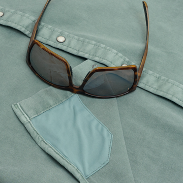 Close-up of "The Ventura" shirt in muted green with a pair of stylish tortoiseshell sunglasses resting on top