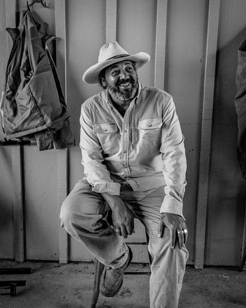 Black and white portrait of a smiling bearded man wearing a hat and button-up shirt on a wooden stool