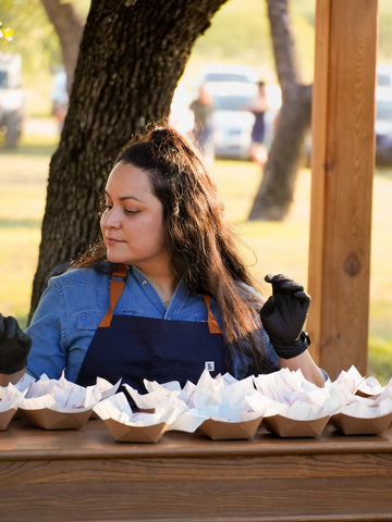 Woman in a blue shirt and apron wearing gloves at an outdoor table with paper food trays