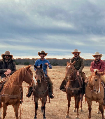 Four horseback riders in cowboy hats and shirts gathered in an open field under cloudy skies