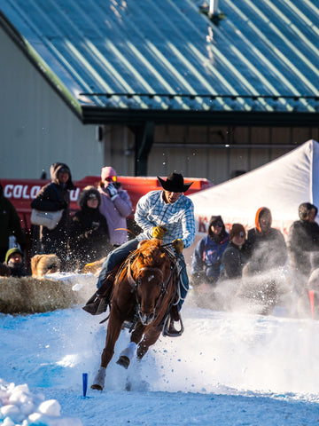 A rider in a plaid shirt and black hat racing a horse through snow with spectators watching behind a barrier.