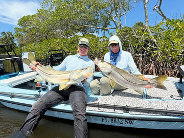 Two anglers hold large fish aboard a boat surrounded by mangroves on a sunny day