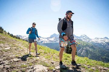 Two hikers trek along a rocky mountain trail with snowy peaks and a sunny alpine landscape behind them