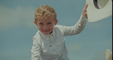 Smiling young boy in white shirt holding a hat outdoors against blue sky