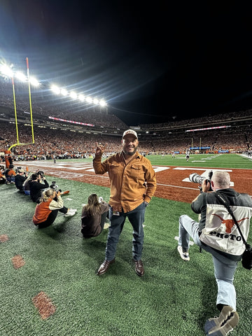 Man in a brown jacket standing on a football field at night with stadium lights and photographers nearby
