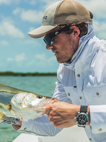 Man wearing Outdoors cap and white fishing shirt holding a fish near water