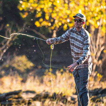 man casts fly while wearing brown and grey plaid long sleeve flannel