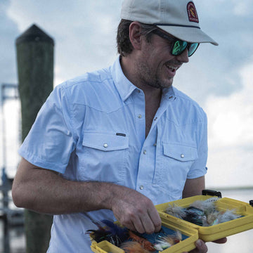 Photo of man with blue microcheck short sleeve shirt standing on dock with a fly box in his hands