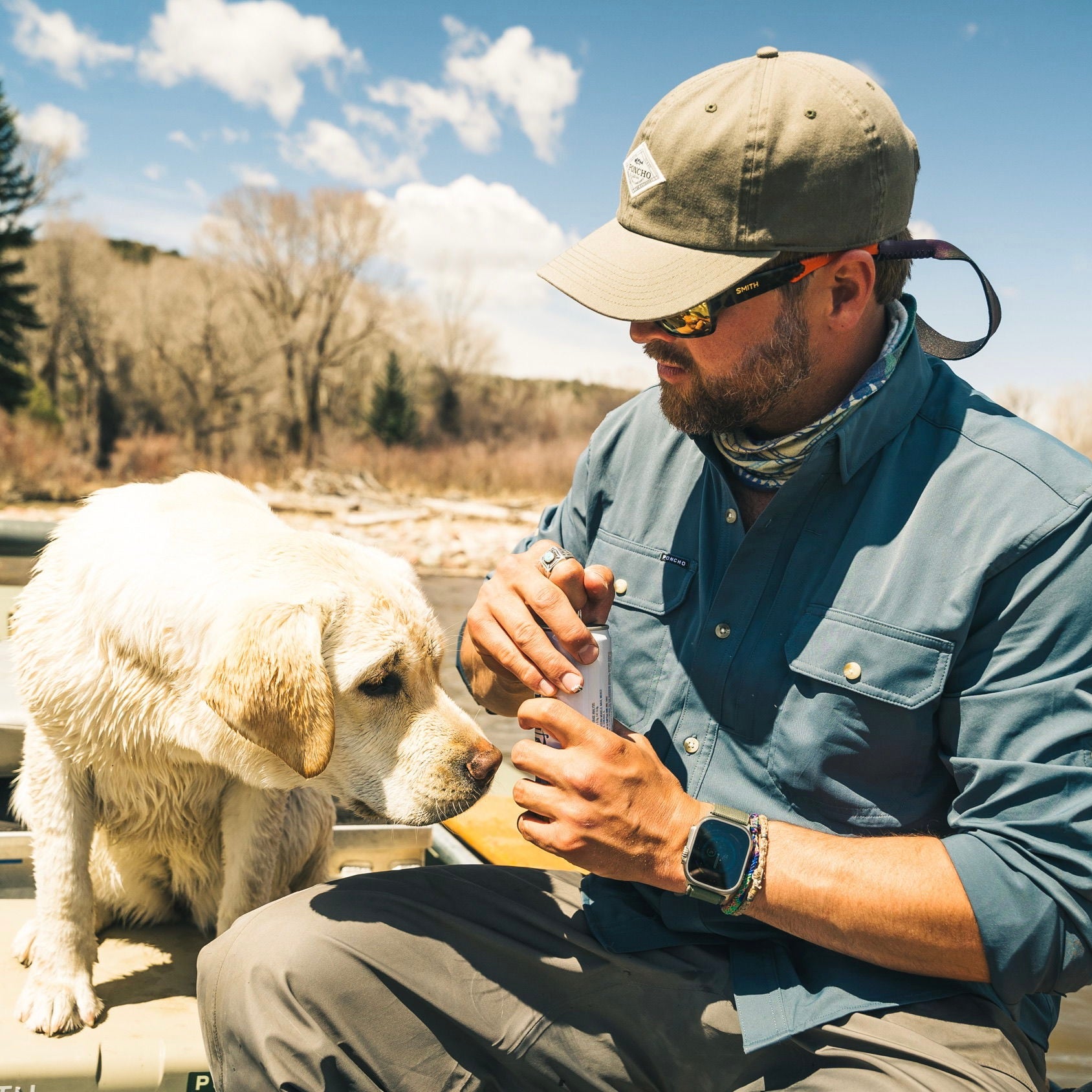 man cracks open a beverage next to curious dog while wearing a deep blue long sleeve fishing shirt