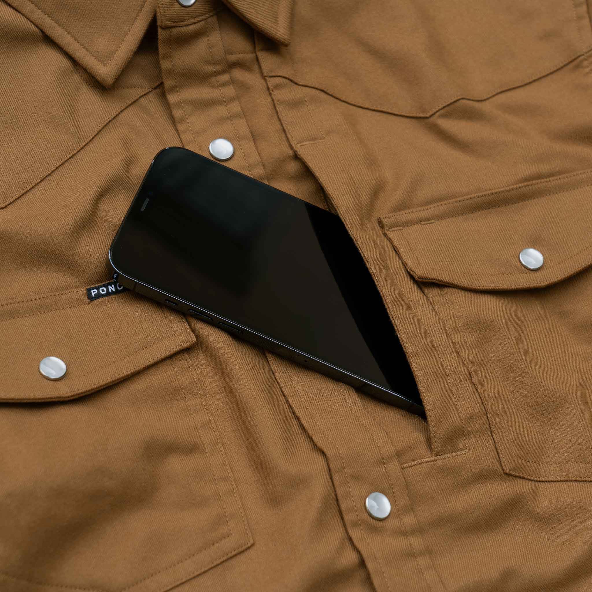 Close up image of a phone going into the chest zipper pocket of the Cheyenne, a tan western flannel