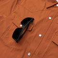 Close up image of sunglasses hanging from chest pocket holder on burnt orange shirt